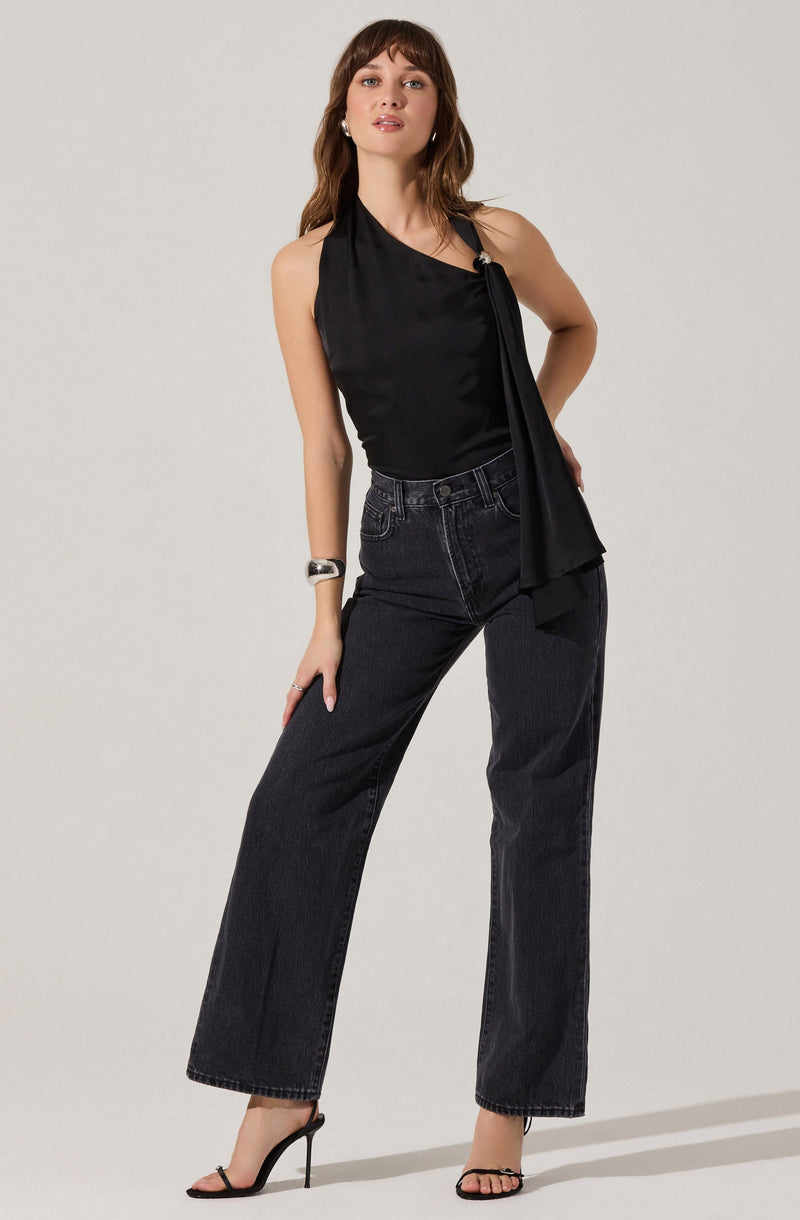 A woman is wearing a sleeveless, asymmetrical black satin top with a large tie detail on the left shoulder. She pairs it with high-waisted, wide-leg dark wash jeans and black high-heeled sandals. She also accessorizes with a chunky silver bracelet on her right wrist.