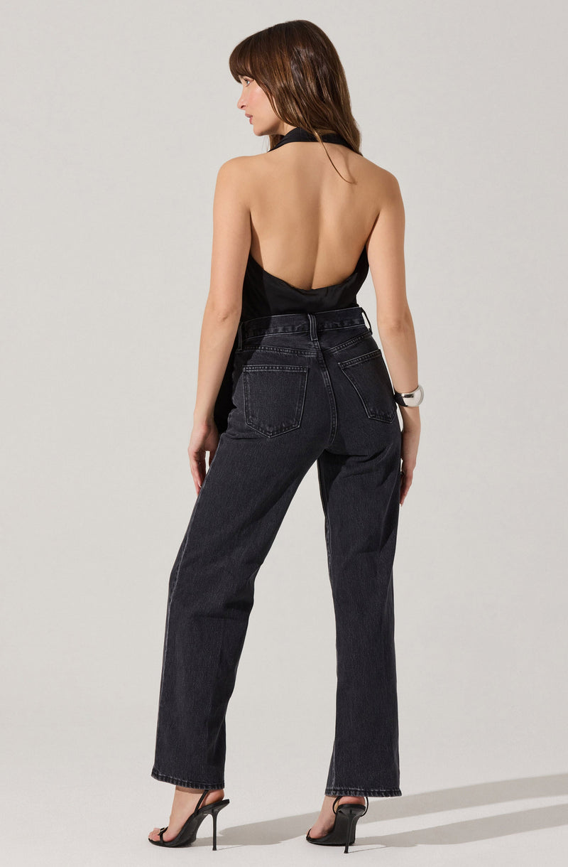 A woman is wearing a black halter-style top with an open back, paired with high-waisted, dark-wash jeans that have a relaxed fit. She is also wearing black high-heeled sandals.