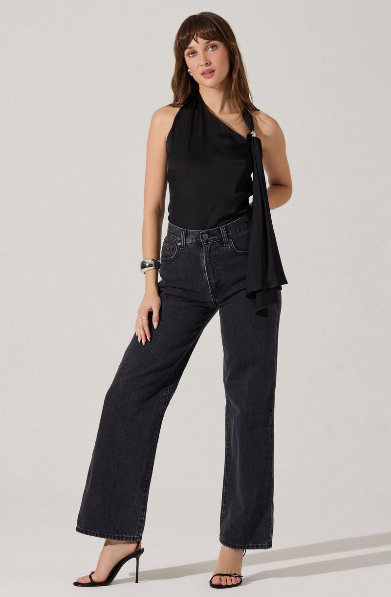 A woman is wearing a sleeveless black satin top with a draped neckline and a large bow detail on the left side. She pairs it with high-waisted, dark wash wide-leg jeans and black high-heeled sandals. She accessorizes with a chunky bracelet on her right wrist and rings on her fingers.