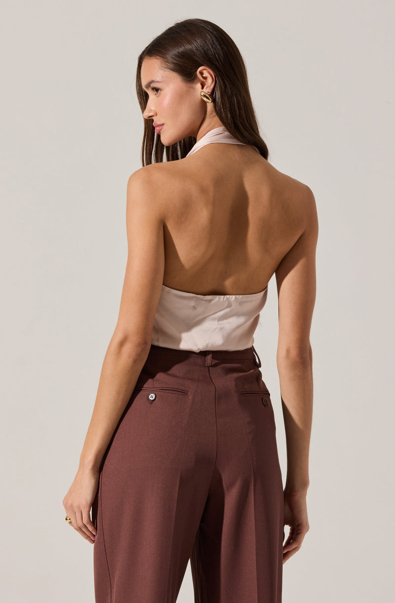 A woman is wearing a sleeveless, high-neck satin top in a light beige color. She pairs it with high-waisted, wide-leg trousers in a deep maroon shade, which feature buttoned welt pockets on the back.