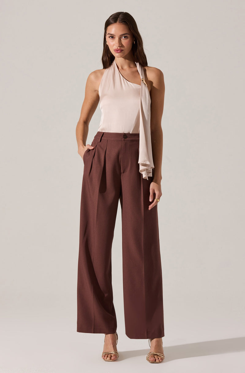 A woman is wearing a sleeveless, light beige blouse with a draped scarf detail hanging from the neckline. She pairs the top with high-waisted, wide-leg brown trousers that feature front pleats and a single button closure. She is also wearing nude, open-toed heels.