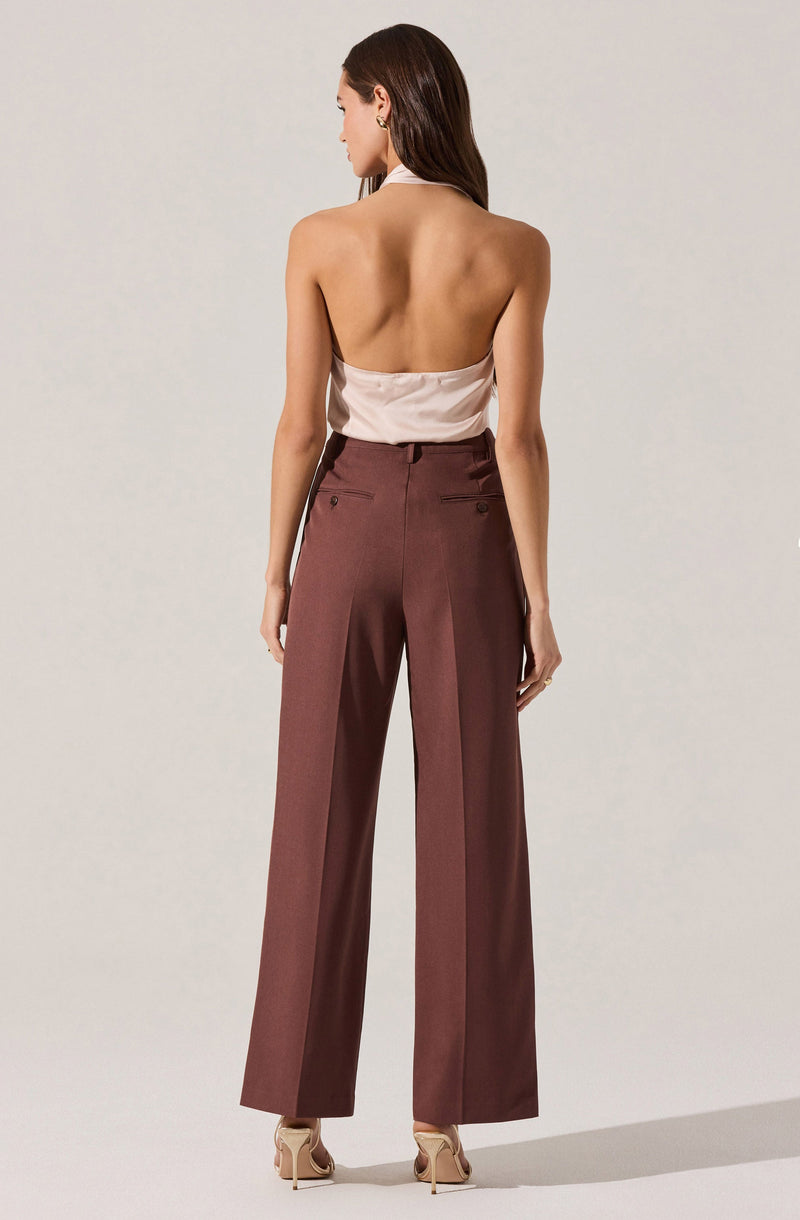 A woman is wearing high-waisted, wide-leg tailored trousers in a deep reddish-brown color. The trousers feature buttoned welt pockets on the back. She is also wearing a light-colored, sleeveless, satin-like top that is tucked into the trousers. The woman has long dark hair and is standing with her back to the camera against a plain, light-colored background.