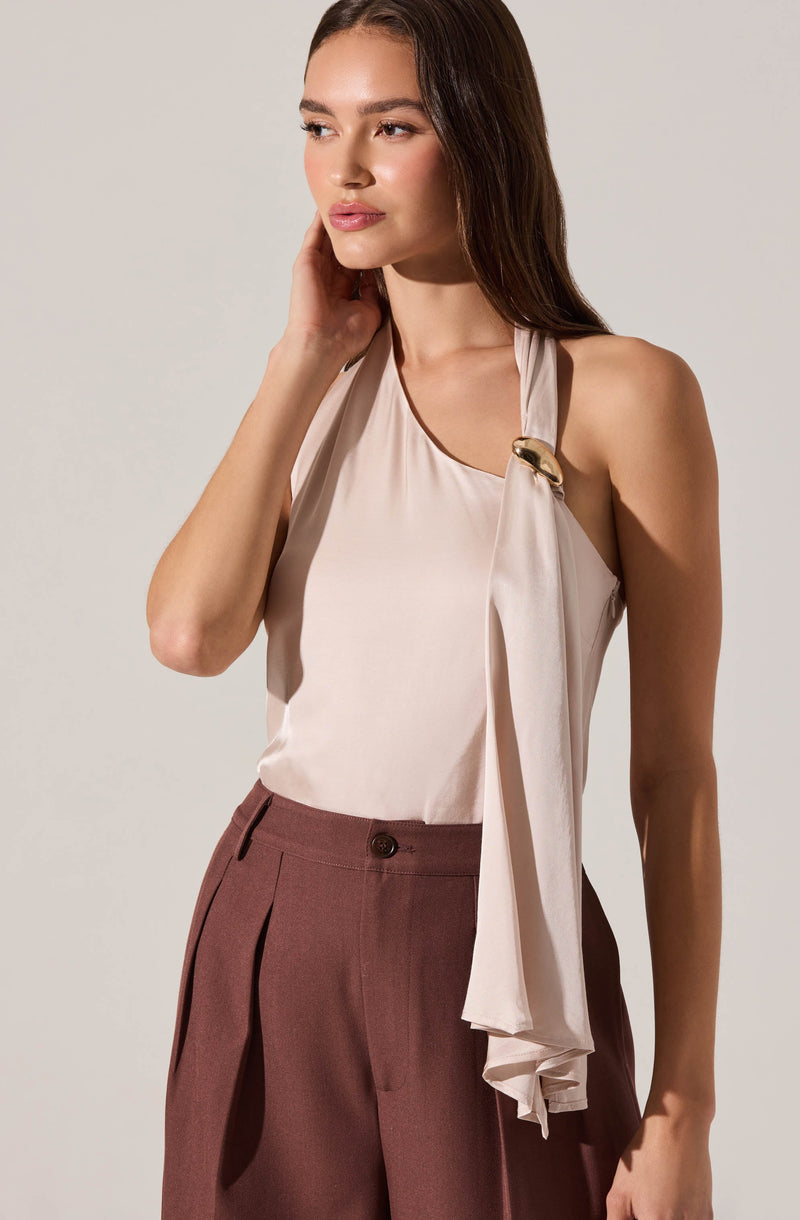 A woman is wearing a sleeveless, cream-colored blouse with a draped tie detail attached at the shoulder, secured with a gold-tone decorative clasp. She pairs it with high-waisted, pleated, brown trousers.