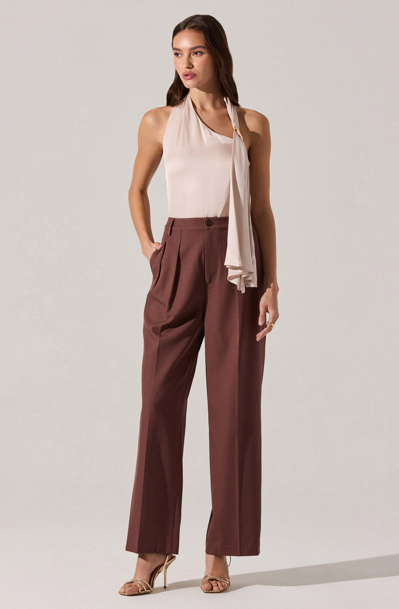 A woman is wearing a sleeveless, light-colored (possibly cream or beige) top with a draped scarf detail hanging from the neckline. She pairs it with high-waisted, wide-leg trousers in a reddish-brown or burgundy color. She completes her outfit with open-toe high-heeled sandals.