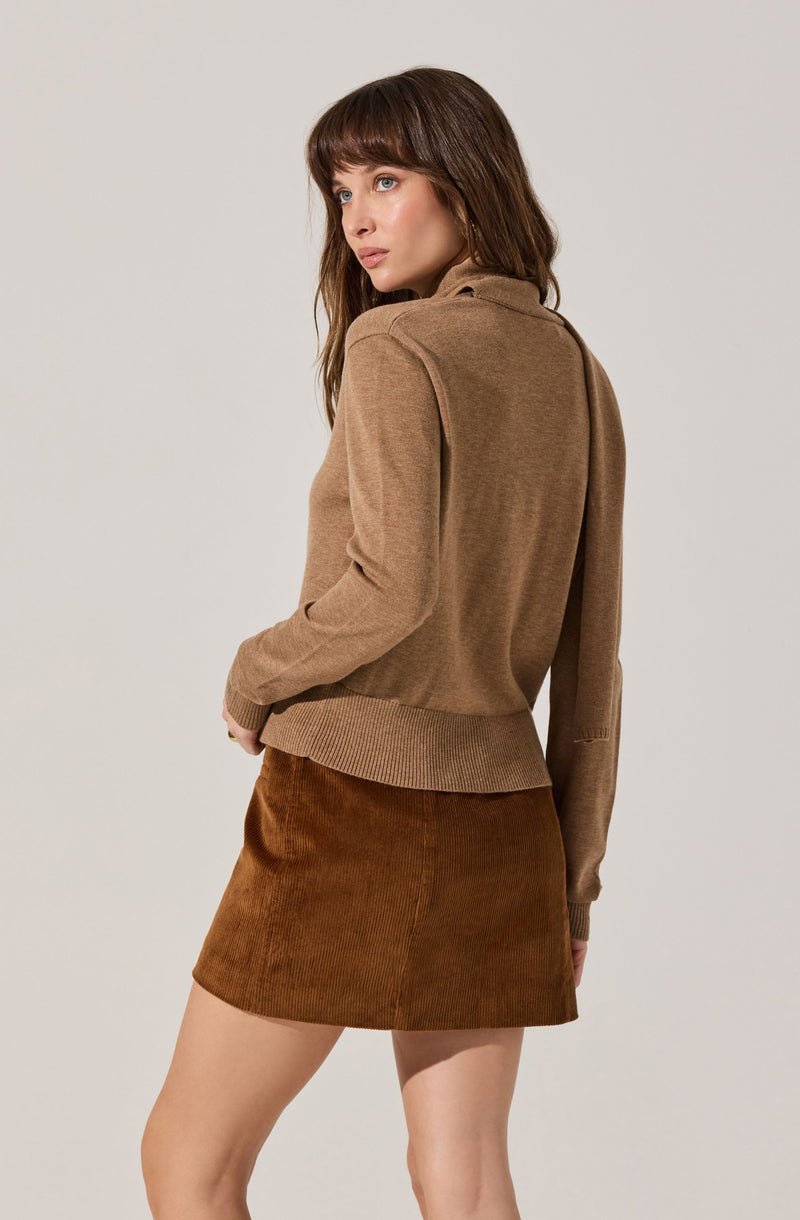 A woman is wearing a light brown, long-sleeved sweater with a high collar and ribbed hem, paired with a brown corduroy skirt. The sweater has a relaxed fit, and the skirt is a mini length.