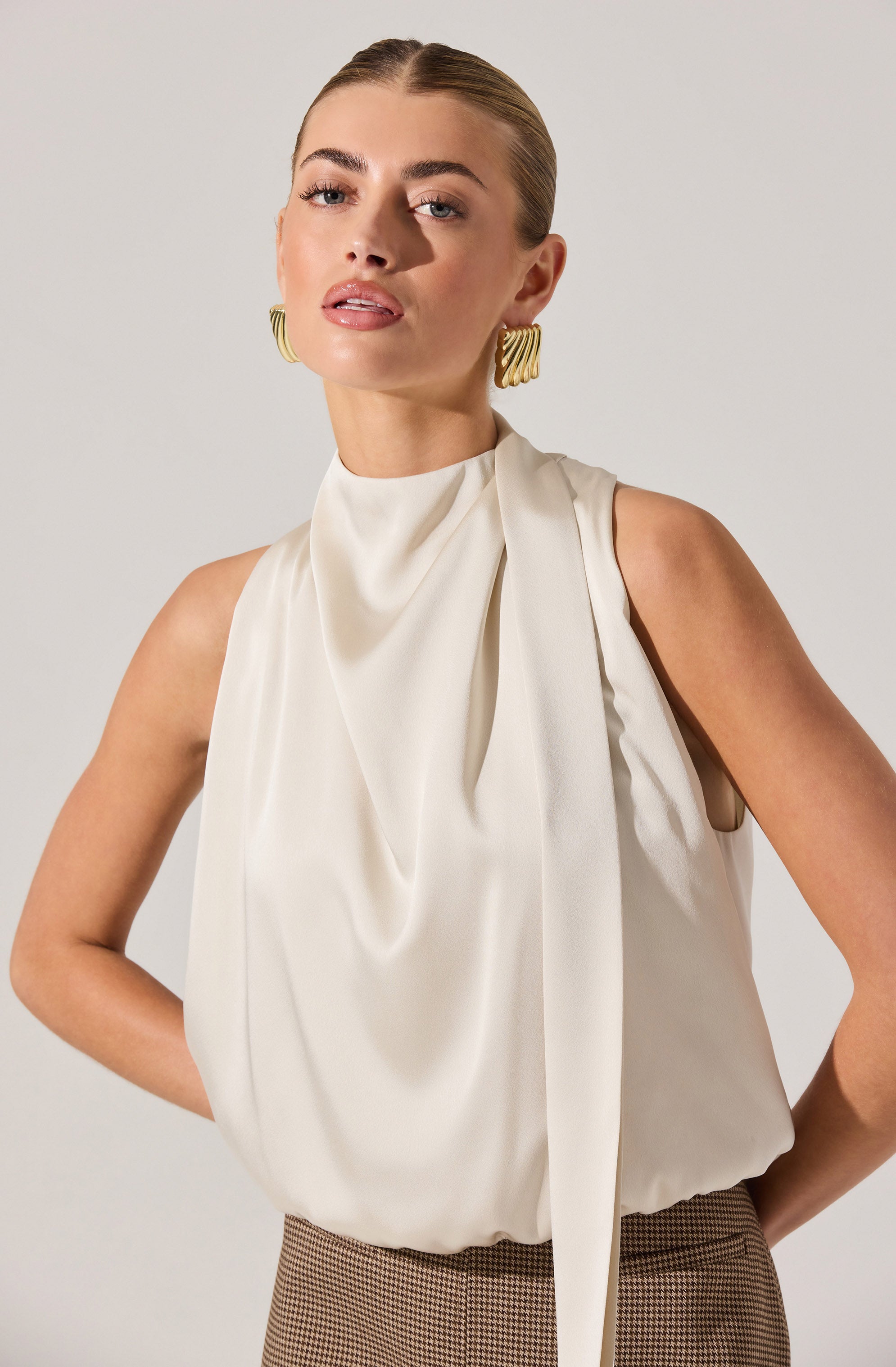 A woman is wearing a sleeveless, ivory-colored blouse with a high neckline and draped fabric details. She has paired the blouse with high-waisted, brown checkered trousers. She is accessorized with large, gold, geometric earrings.