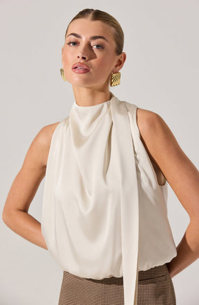 A woman is wearing a sleeveless, ivory-colored blouse with a high neckline and draped fabric details. She has paired the blouse with high-waisted, brown checkered trousers. She is accessorized with large, gold, geometric earrings.