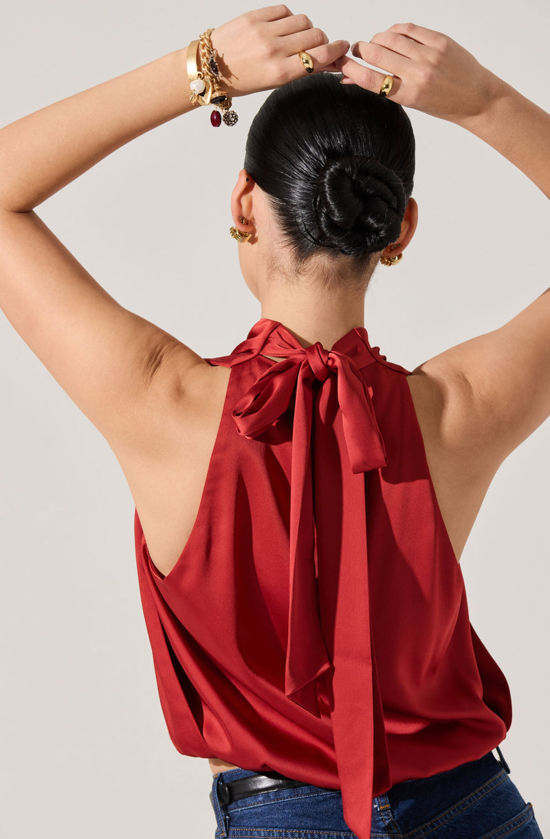 A woman is wearing a sleeveless red satin top with a large bow tied at the back of the neckline. She is also wearing dark blue jeans. Her hair is styled in a sleek, low bun, and she has multiple gold earrings and bracelets, some adorned with charms and stones.