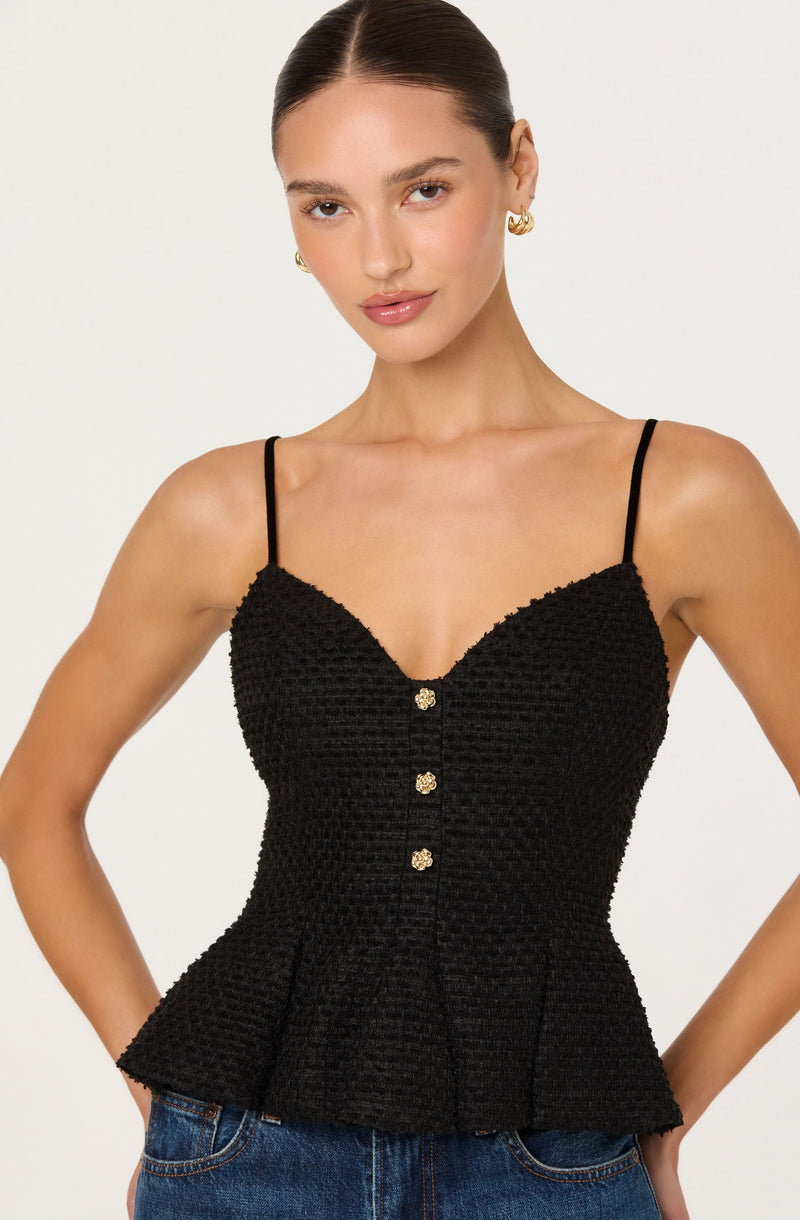 A woman is wearing a black sleeveless top with thin spaghetti straps. The top features a textured fabric with a peplum hem and decorative buttons down the front, each adorned with small, shiny embellishments.