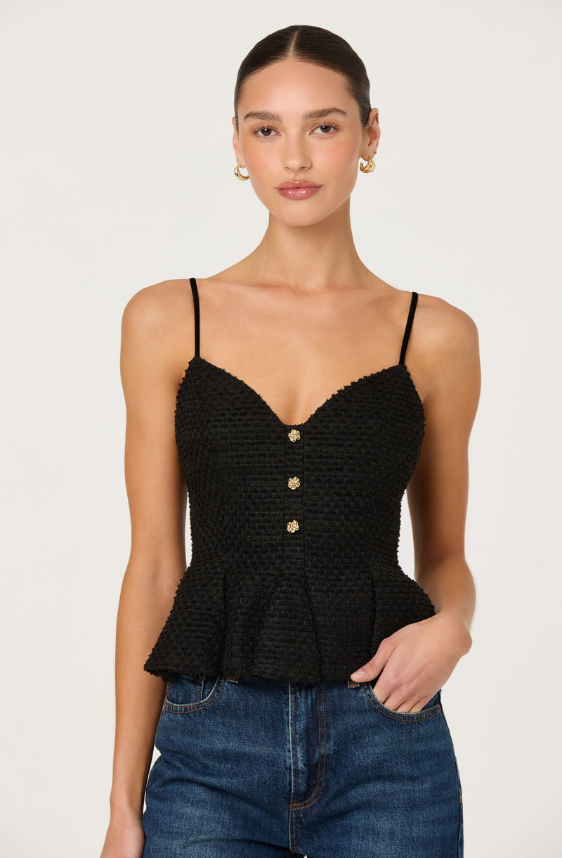 A woman is wearing a black, textured spaghetti strap top with a fitted waist and a slight peplum hem. The top features three decorative buttons down the front. She is also wearing blue denim jeans and gold hoop earrings.