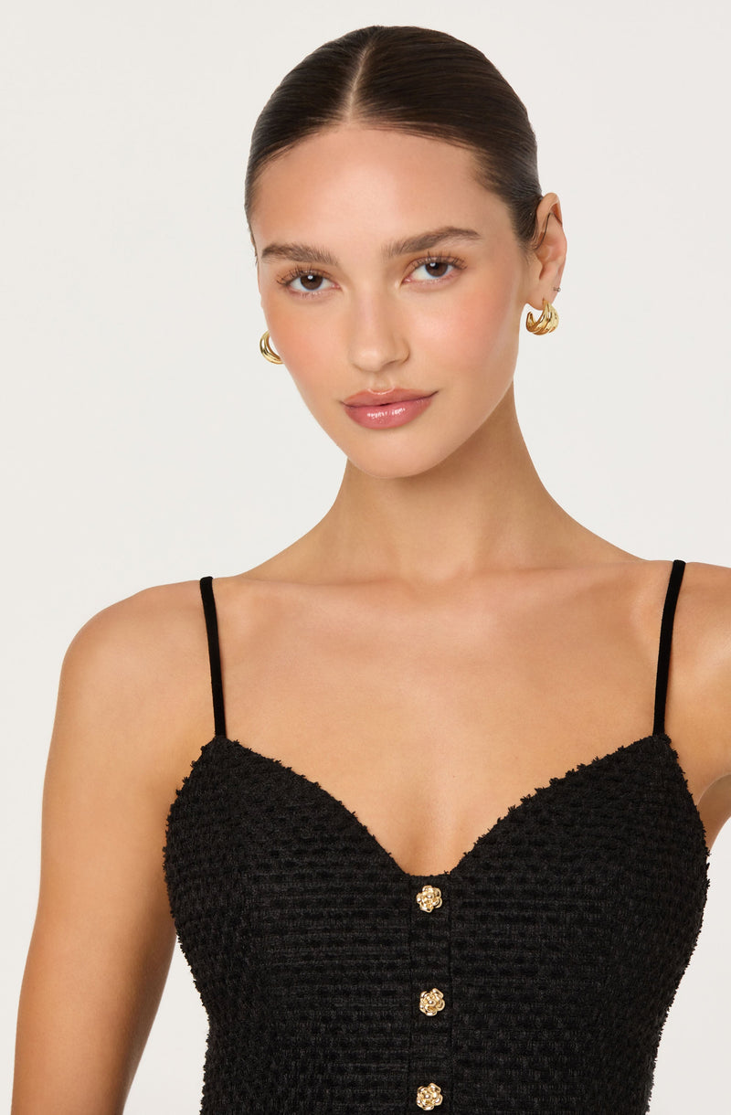 A woman is wearing a black textured spaghetti strap top with thin straps and a row of decorative buttons down the front. She is also wearing gold hoop earrings.
