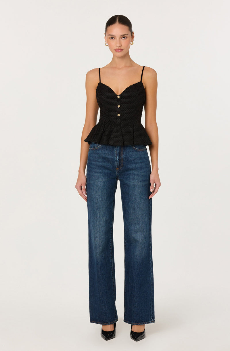A woman is wearing a black, textured, sleeveless top with thin spaghetti straps and decorative buttons down the front. The top has a peplum style with a ruffled hem. She is also wearing high-waisted, wide-leg blue jeans and black pointed-toe shoes.