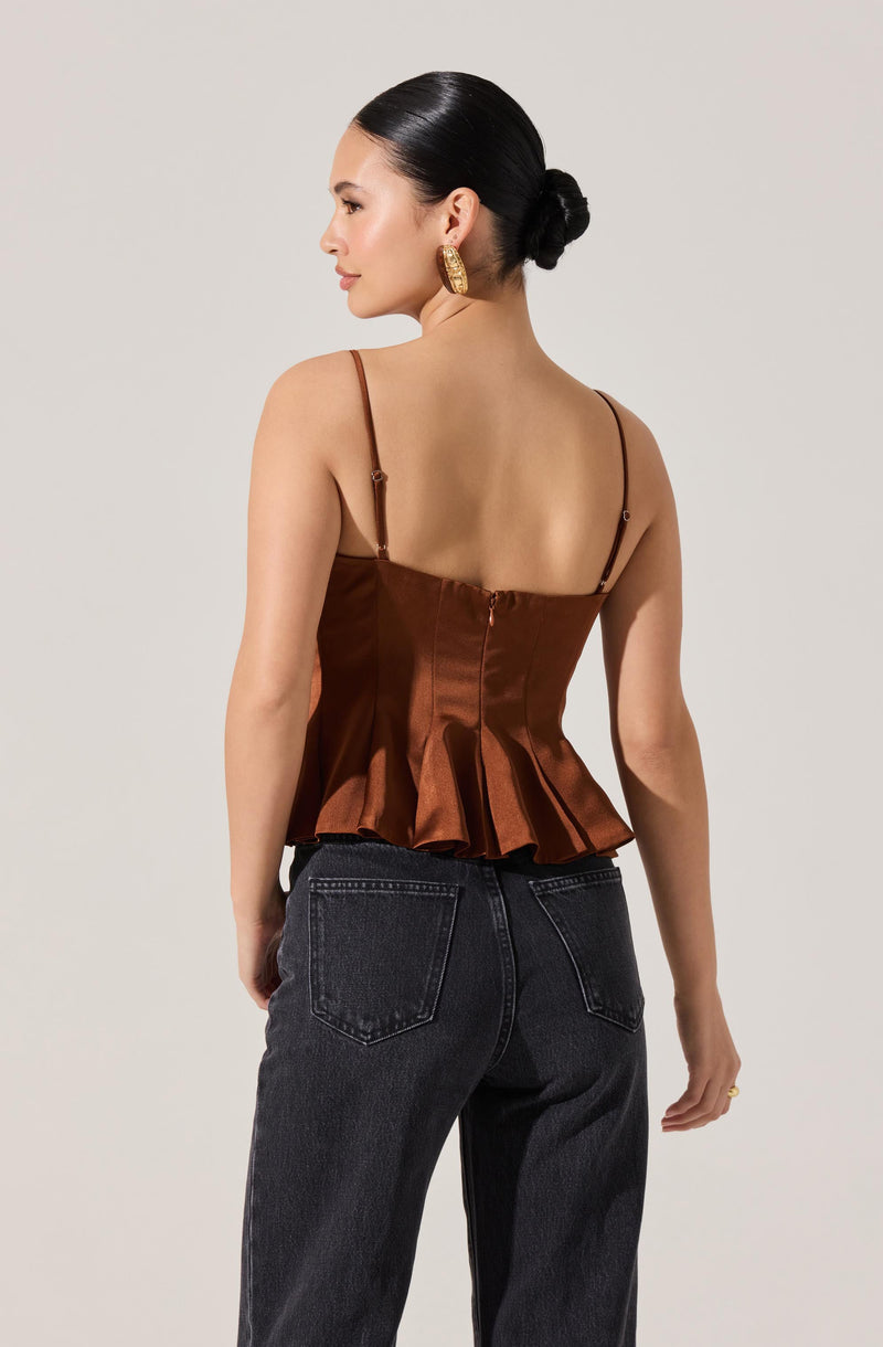 A woman is wearing a bronze satin sleeveless top with thin shoulder straps, a fitted bodice, and a peplum hem. She pairs it with high-waisted, dark-wash jeans with visible back pockets. She has her hair styled in a low bun and is wearing large, gold hoop earrings.
