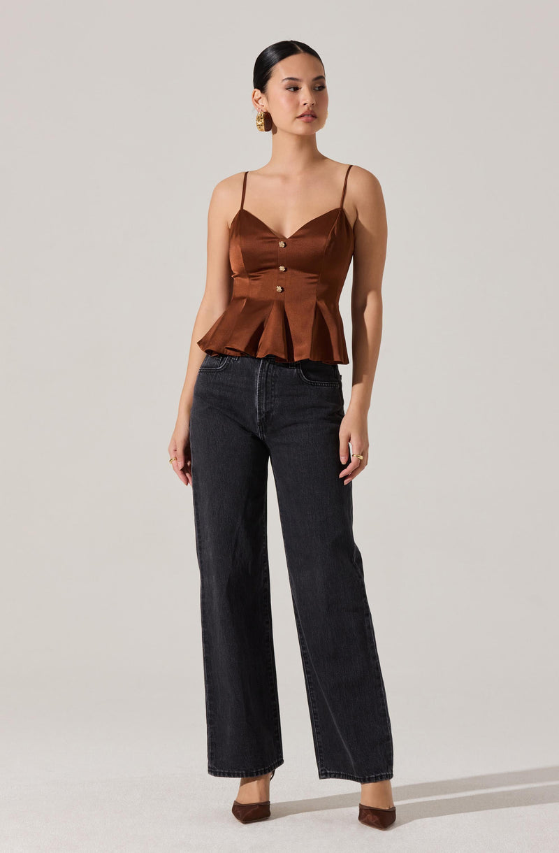 A woman is wearing a brown satin sleeveless top with thin spaghetti straps, button details down the front, and a peplum flare at the hem. She pairs it with high-waisted, straight-leg black jeans and brown pointed-toe heels. She also has gold earrings and rings on her fingers.
