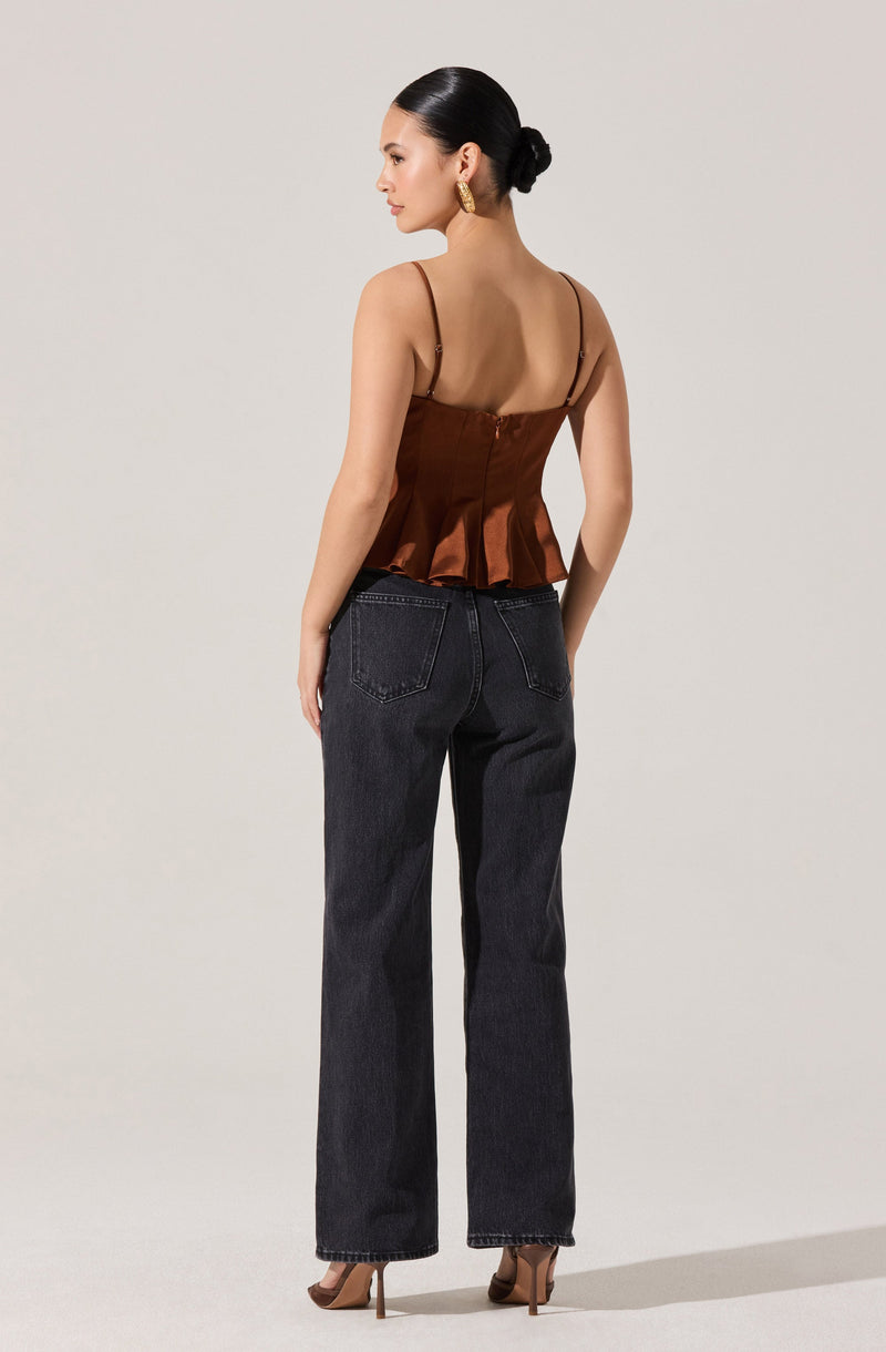 A woman is wearing a sleeveless brown satin top with thin spaghetti straps and a ruffled hem, paired with high-waisted, straight-leg black jeans. She has her hair styled in a low bun and is accessorizing with large gold earrings.