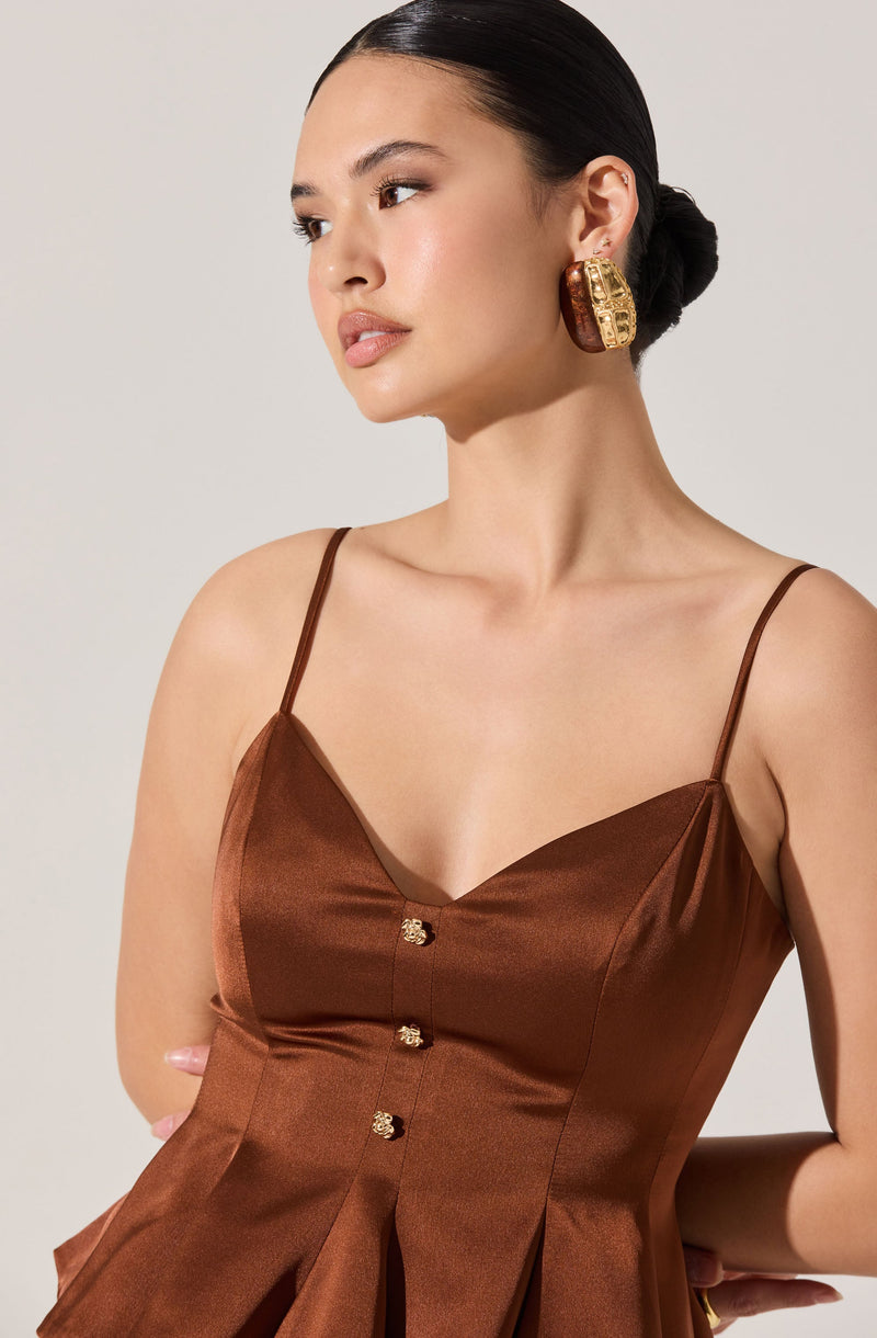 A woman is wearing a sleeveless, brown satin dress with thin spaghetti straps. The dress has a fitted bodice with a v-neckline and decorative buttons down the front. The fabric appears smooth and shiny, with a slightly gathered waist creating a subtle flare at the skirt. She is also wearing large, gold and brown statement earrings.