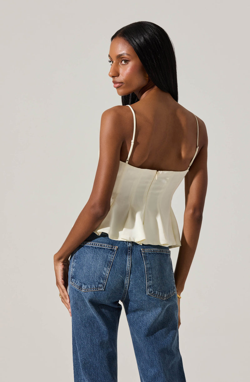 A woman with dark hair styled straight is wearing a cream-colored, sleeveless, peplum top with thin spaghetti straps and a back zipper. She is also wearing high-waisted blue jeans with visible stitching and back pockets.