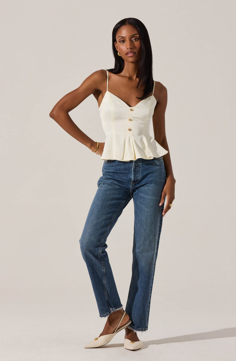 A woman is wearing a white sleeveless top with thin spaghetti straps, featuring a V-neckline and button details down the front, along with a flared hem. She pairs it with high-waisted, straight-leg blue jeans and beige pointed-toe heels.