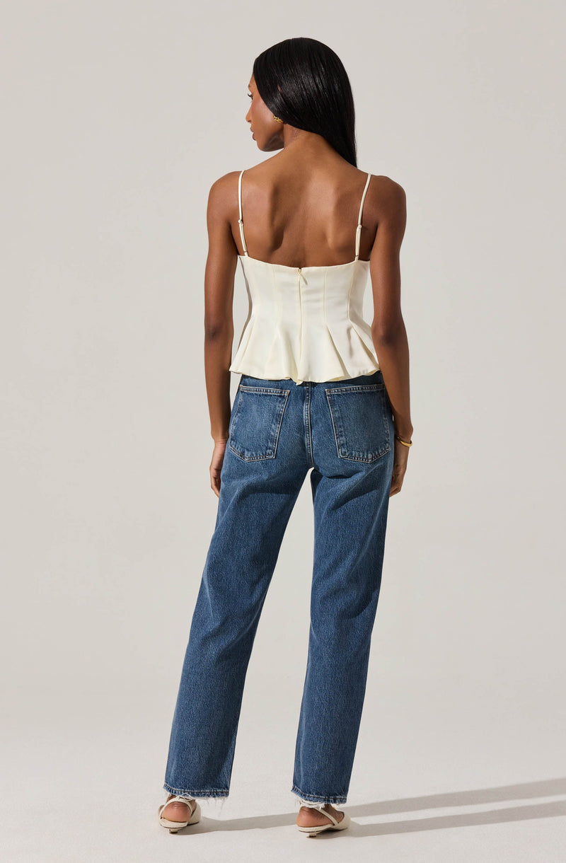 A woman is wearing a cream-colored, sleeveless, corset-style top with thin spaghetti straps and a peplum hem. She has on high-waisted, loose-fitting blue jeans with visible back pockets, and nude-colored heeled sandals.