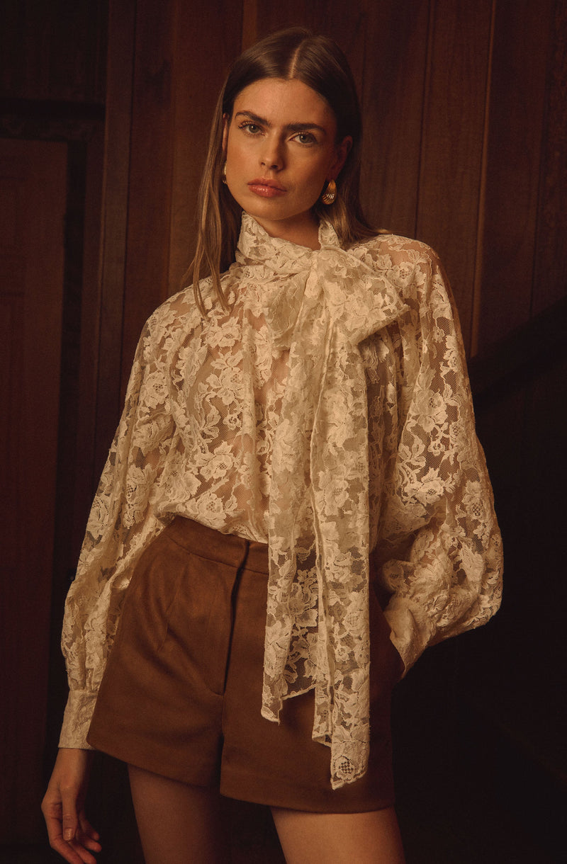 A woman is wearing a cream-colored lace blouse with a large bow tied at the neck, featuring long, slightly puffed sleeves. She pairs it with high-waisted brown shorts. The background consists of dark wooden paneling.