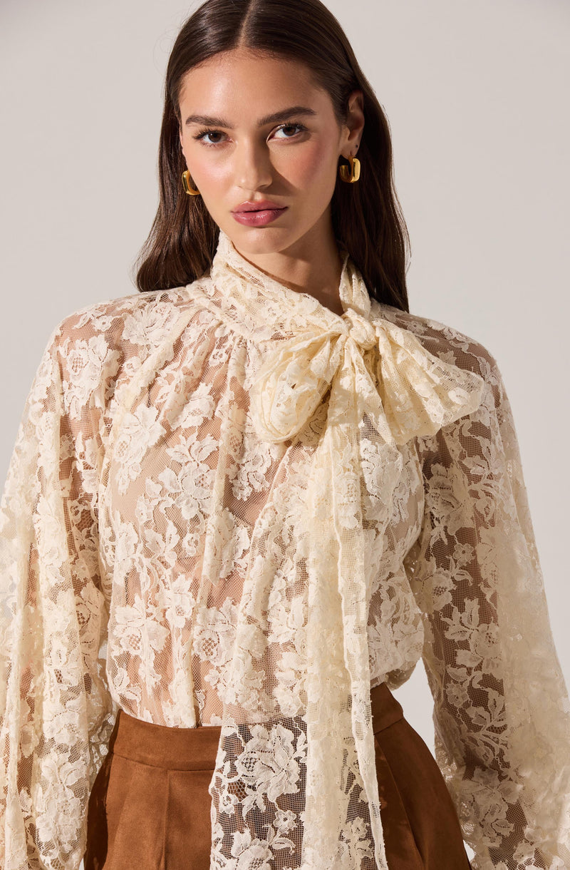 A woman is wearing a long-sleeved, cream-colored lace blouse with a large bow at the neck. The blouse features a floral lace pattern with sheer details. She has paired the blouse with high-waisted brown trousers. She accessorizes with gold hoop earrings.