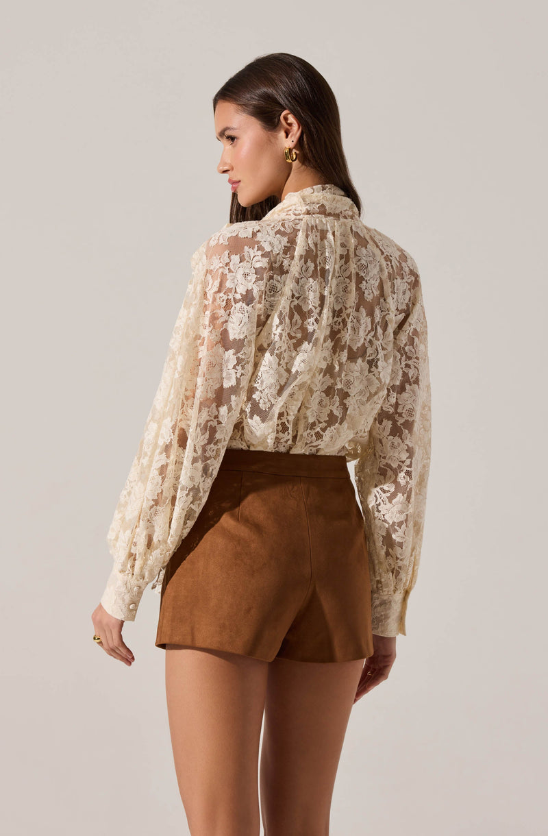 A woman is wearing a long-sleeved, cream-colored lace blouse with a high neckline and gathered detail at the shoulders, paired with high-waisted, brown suede shorts that have a tailored fit.