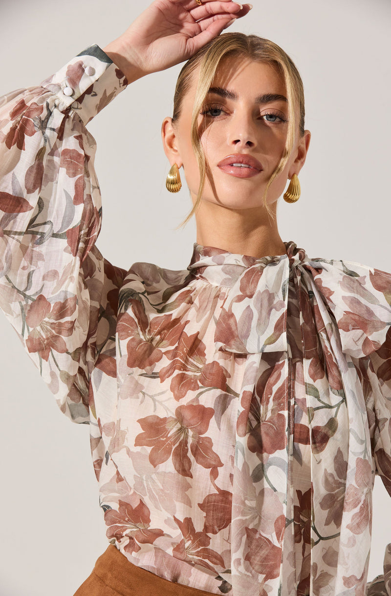 A woman is wearing a long-sleeved, semi-sheer blouse with a floral pattern in shades of pink, brown, and beige. The blouse has a high neckline with a large bow tie detail at the front. She is also wearing gold, textured hoop earrings.