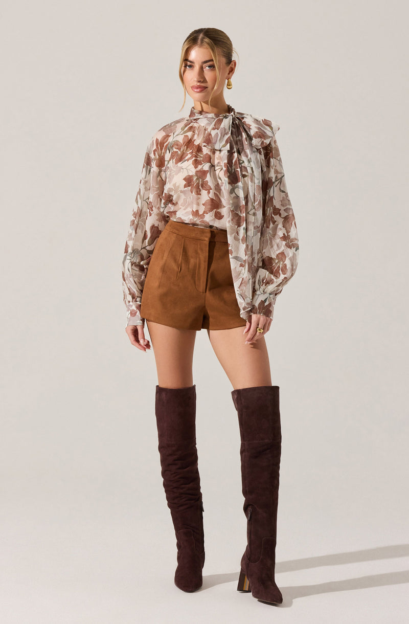 A woman is wearing a long-sleeved blouse with a floral print in shades of beige, brown, and cream, featuring a large bow tied at the neck. She pairs it with high-waisted brown shorts and tall, dark brown suede over-the-knee boots with a heel.