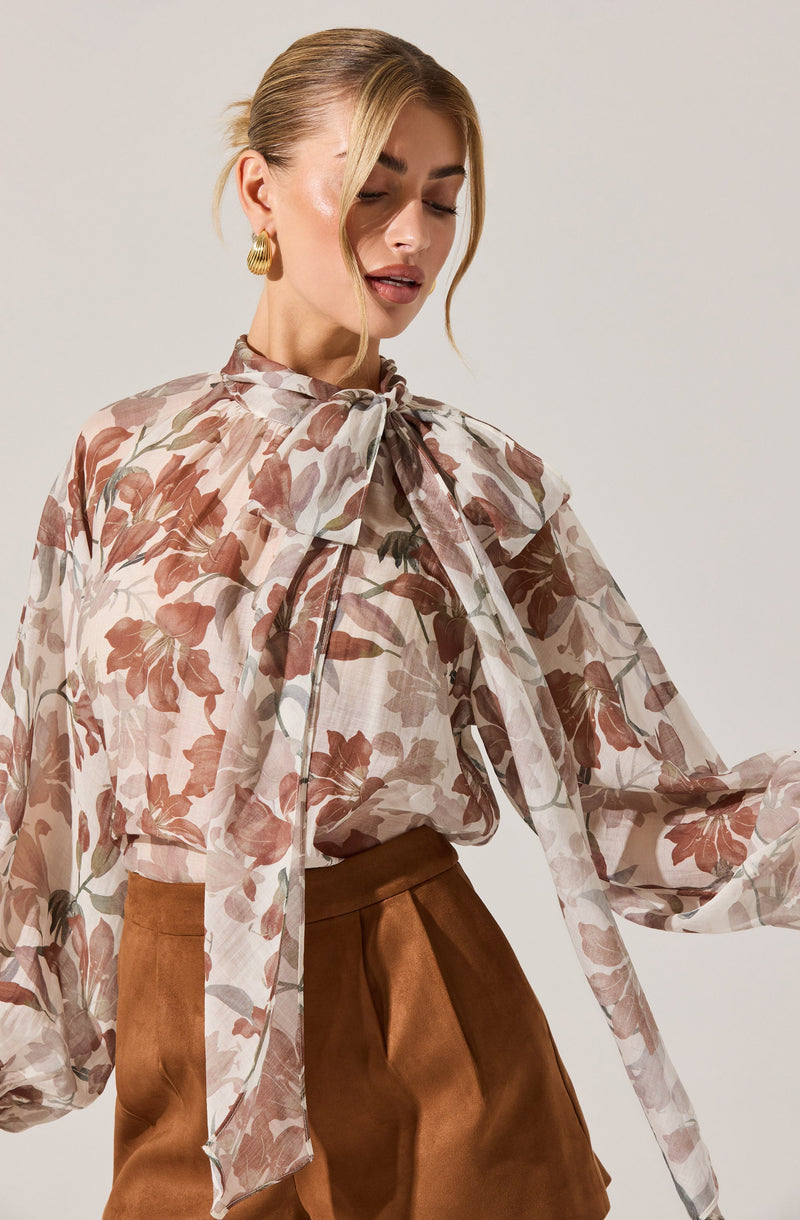 The woman is wearing a sheer, long-sleeved blouse with a large bow at the neckline. The blouse features a floral pattern in shades of brown, beige, and muted pink. She pairs it with high-waisted, brown satin-style pants.
