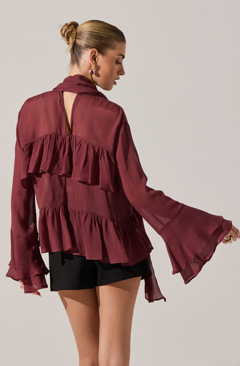 A woman is wearing a sheer, long-sleeved, maroon blouse with ruffle details on the back and sleeves. The blouse features a keyhole opening at the back neckline. She pairs it with black shorts. She is also wearing earrings, and her hair is styled in a bun.