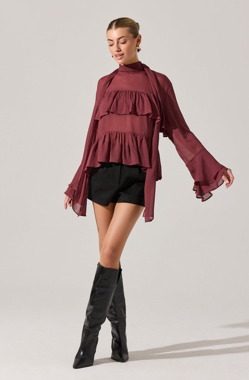 A woman is wearing a long-sleeved, high-neck, burgundy blouse with layered ruffles on the front and flowy, sheer sleeves. She pairs it with black shorts and tall, black leather boots.