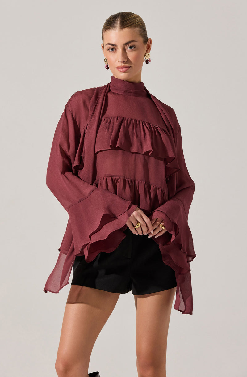 A woman is wearing a high-neck, long-sleeved blouse in a deep burgundy color with layered ruffles across the front and on the flared, voluminous sleeves. She pairs it with black shorts. She has her hair pulled back and is accessorized with statement earrings and rings.