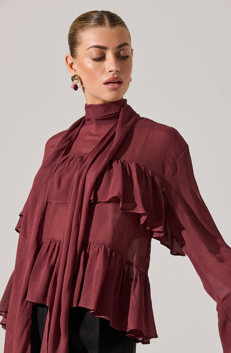 The woman is wearing a long-sleeved, deep burgundy blouse with ruffled details on the front and sleeves. The blouse features a high neckline with a matching tie or scarf-like extension hanging down the front. She has paired it with large, colorful earrings.