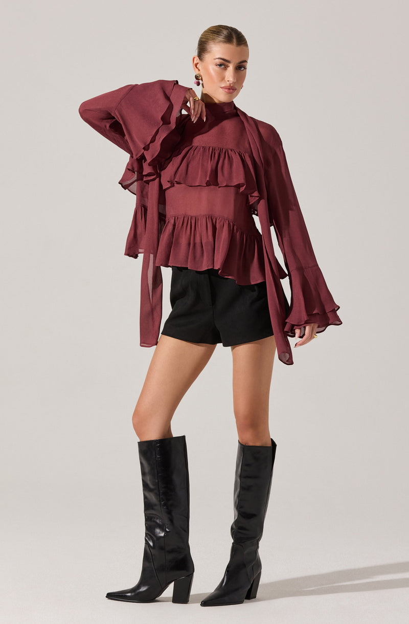 A woman is wearing a long-sleeved, ruffled, burgundy blouse with layered ruffles across the chest and waist, and wide, ruffled cuffs on the sleeves. She pairs it with black shorts and tall, shiny black knee-high boots with pointed toes and high block heels.