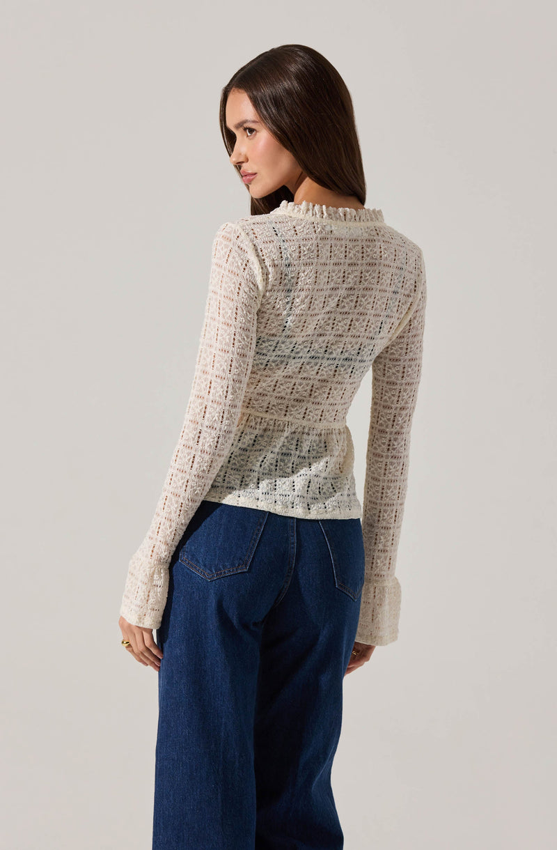 A woman is wearing a long-sleeved, cream-colored, knit top with a square-patterned lace design. The top has a ruffled neckline and cuffs. She is also wearing high-waisted, dark blue jeans with visible back pockets.
