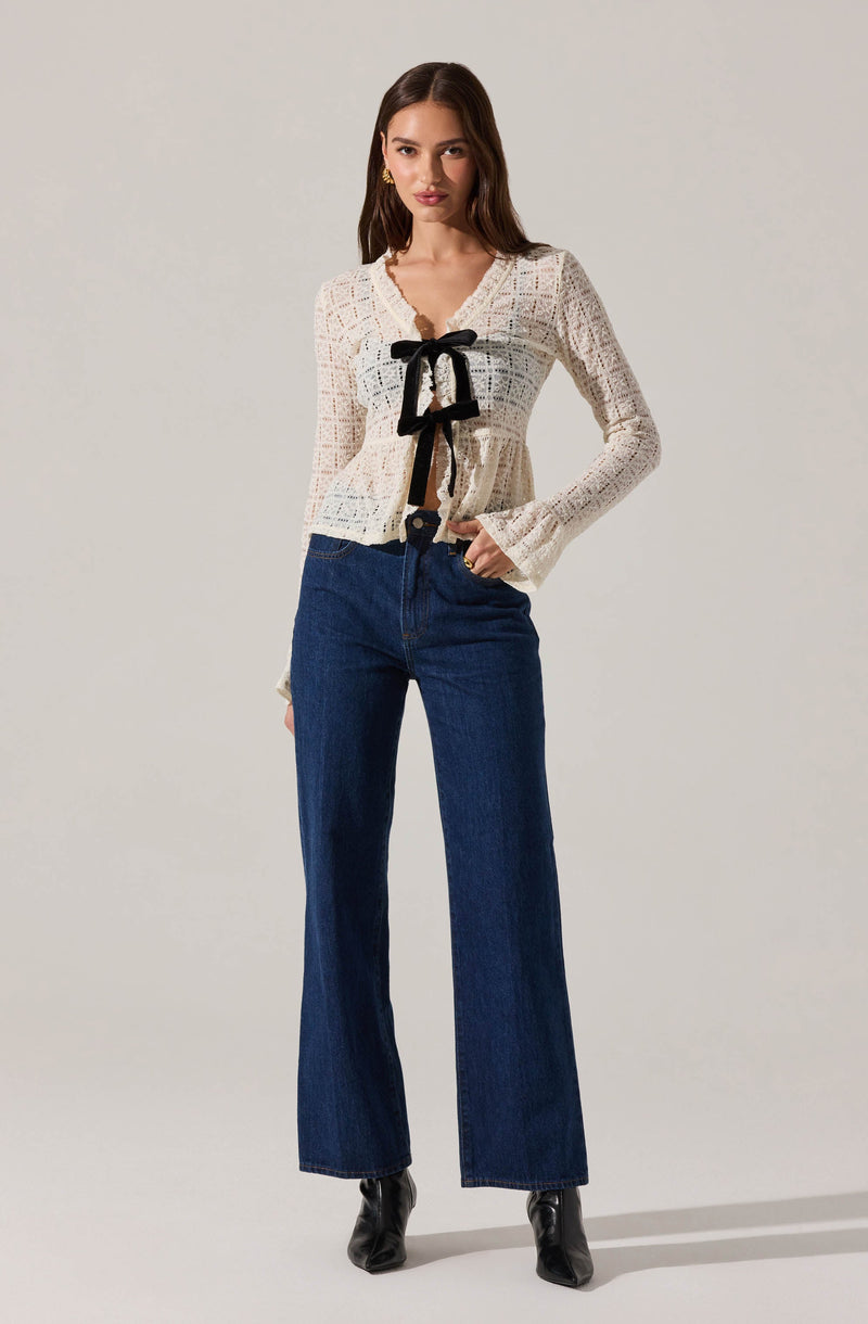 A woman is wearing a white, long-sleeved crochet top with a scalloped hem and sleeves, featuring a black bow tie detail at the neckline with matching black ribbon accents. She pairs the top with high-waisted, dark blue wide-leg jeans and black pointed-toe ankle boots.
