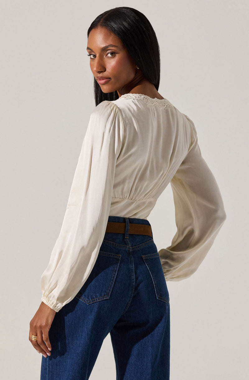 A woman with long, straight black hair is wearing a cream-colored, long-sleeved blouse with a subtle sheen. The blouse has a gathered elastic waist and delicate eyelet lace detail along the neckline. She pairs it with high-waisted dark blue jeans and a brown belt.
