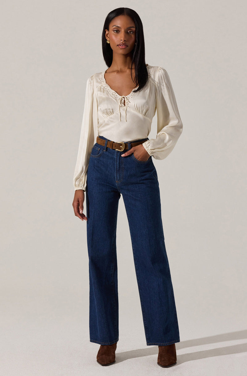 A woman is wearing a long-sleeved, cream-colored blouse with lace detailing around the neckline and a small tie at the front. She has paired it with high-waisted, dark-wash wide-leg jeans and a brown belt. She is also wearing brown boots.