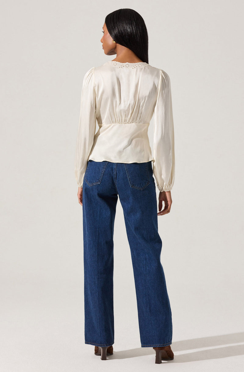 A woman is wearing a long-sleeved, cream-colored blouse with a subtle sheen and gathered waist, featuring delicate eyelet embroidery along the neckline. She is paired with high-waisted, straight-leg blue jeans and brown heeled shoes.