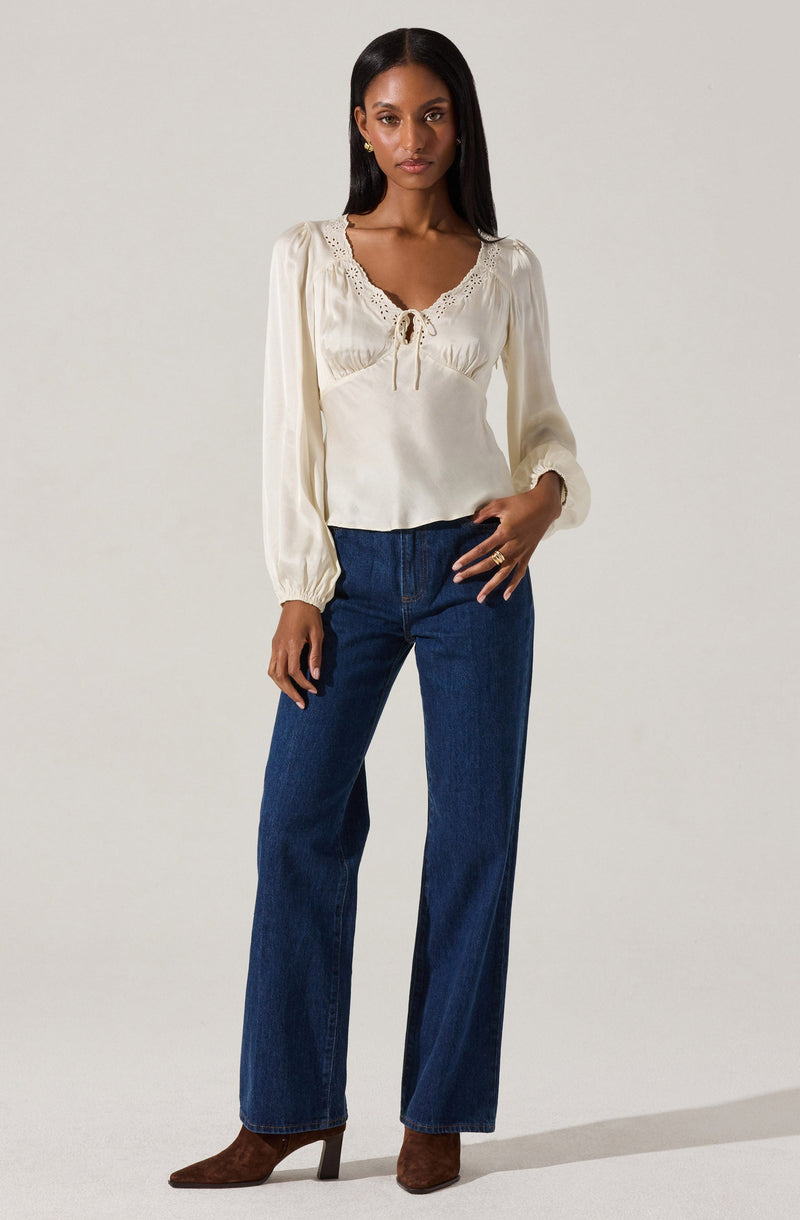 A woman is wearing a long-sleeved, cream-colored blouse with a lace trim around the neckline and a small bow at the center. She is also wearing high-waisted, dark blue jeans with a wide-leg fit and brown heeled boots.