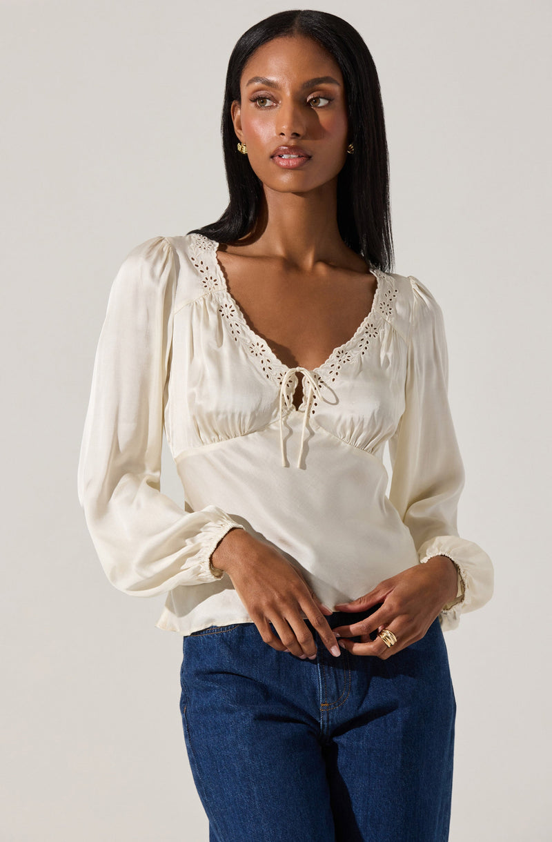 A woman is wearing a long-sleeved, cream-colored blouse with a lace trim around the neckline and a small bow tie at the front. The blouse has puffed shoulders and gathered cuffs. She is also dressed in dark blue jeans.