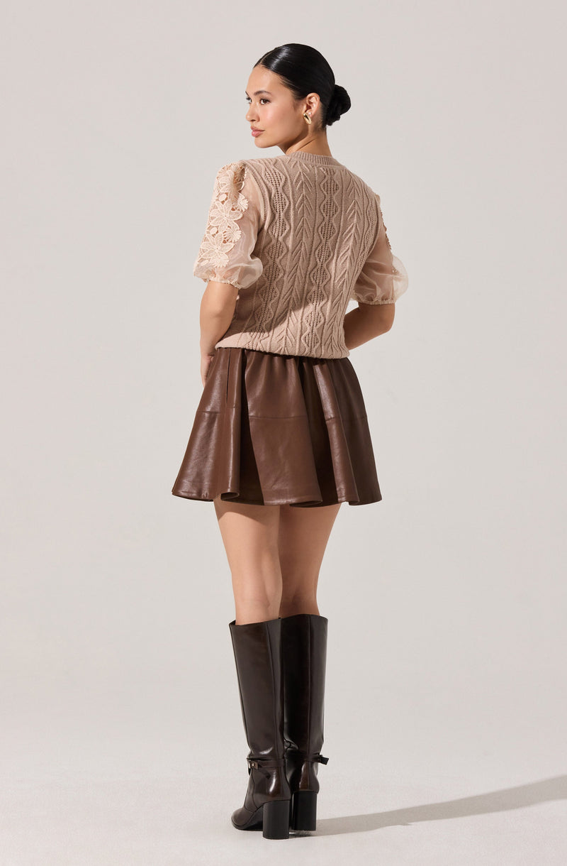 A woman is wearing a beige cable-knit sweater with floral lace sleeves, paired with a brown leather flared skirt. She completes the outfit with tall black leather boots and has her dark hair styled in a low bun.