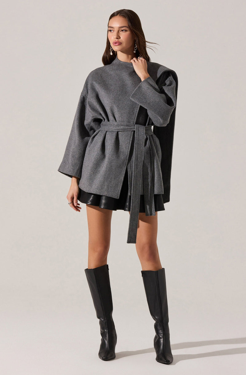 A woman is wearing a gray oversized coat with long sleeves and a belt at the waist. The coat has a darker gray panel on one shoulder, adding contrast. She pairs the coat with black leather shorts and knee-high black leather boots.
