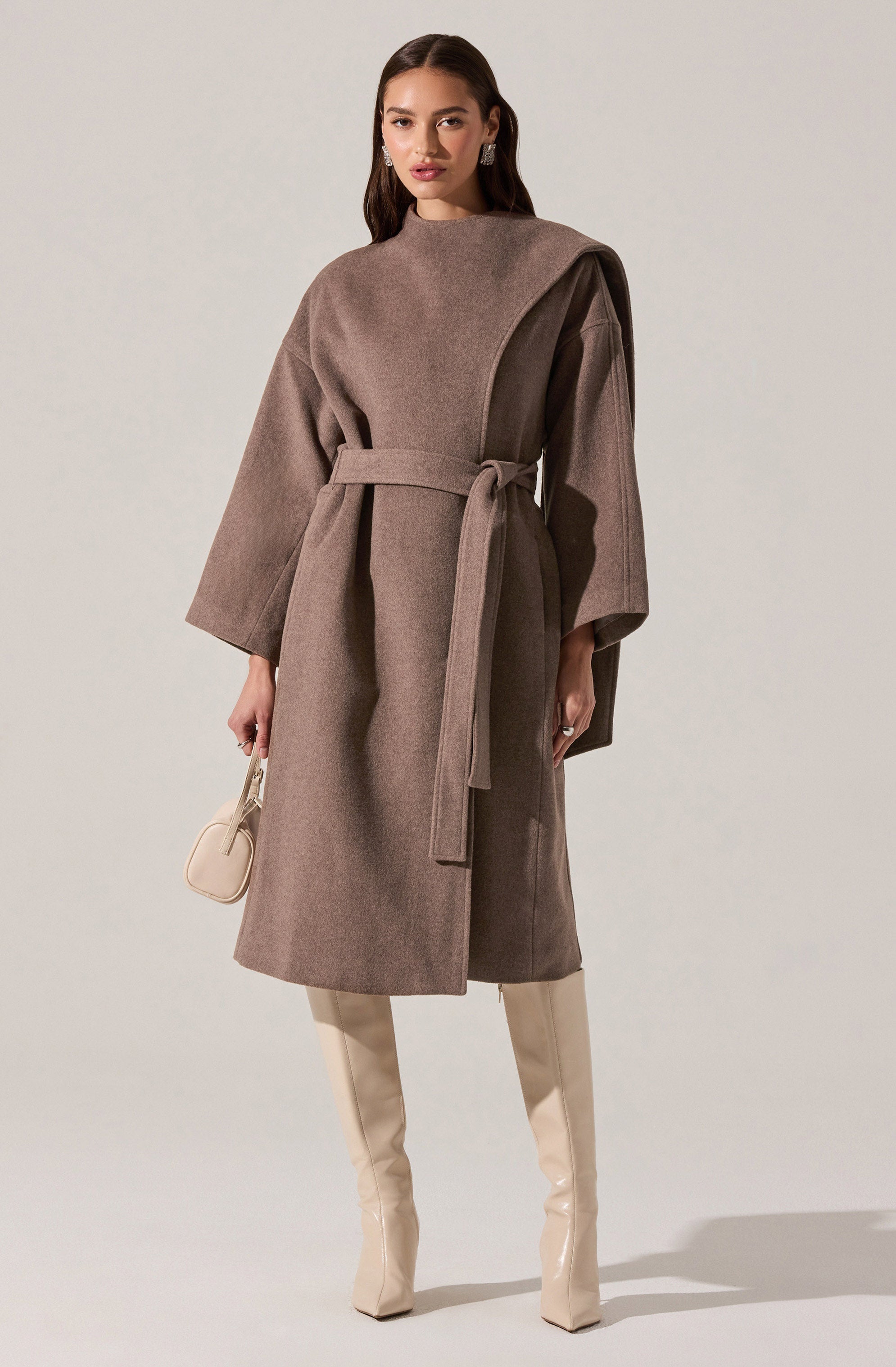 A woman is wearing a long, taupe-colored coat with wide sleeves and a matching belt tied at the waist. The coat features a large, asymmetrical shoulder detail on her left side. She is also wearing high, cream-colored boots and holding a small, cream-colored handbag. She has dark, wavy hair and is accessorized with earrings.