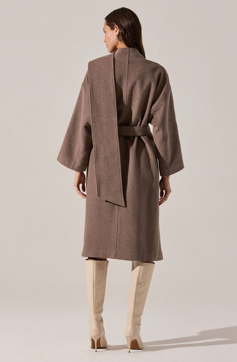A woman is wearing a long, taupe-colored coat with wide sleeves and a matching belt tied at the waist. She has a scarf draped over her shoulders that matches the coat’s color. She is also wearing tall, cream-colored high-heeled boots.