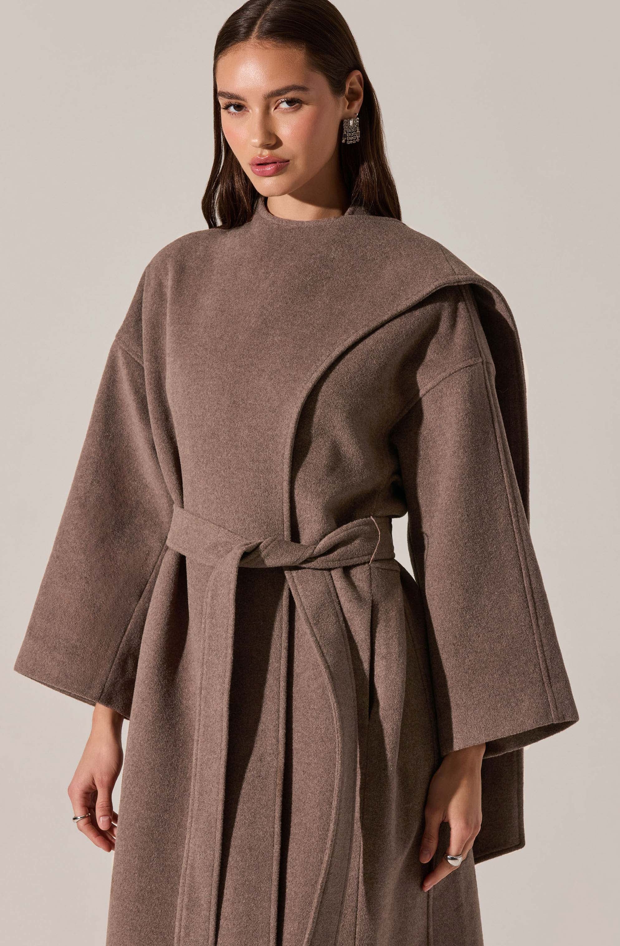 A woman is wearing a taupe-colored, oversized coat with wide sleeves. The coat features a large, draped collar and a matching fabric belt tied at the waist. The fabric appears to be a soft, wool or wool-blend material.