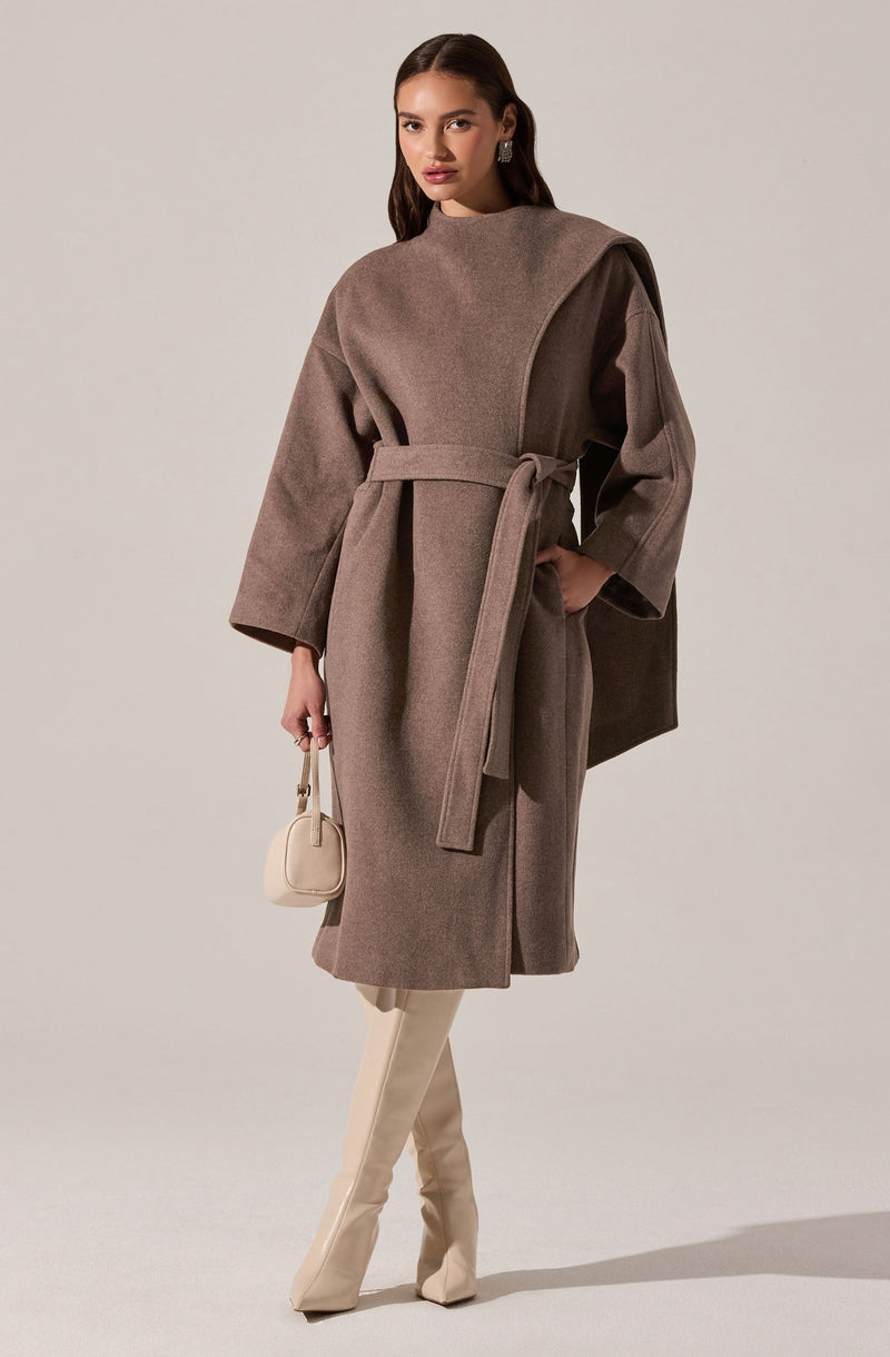 A woman is wearing a long taupe-colored coat with wide sleeves and a matching belt tied around her waist. The coat has a high neckline and a draped, asymmetrical design on one shoulder. She is holding a small, cream-colored handbag and is wearing cream-colored knee-high boots.