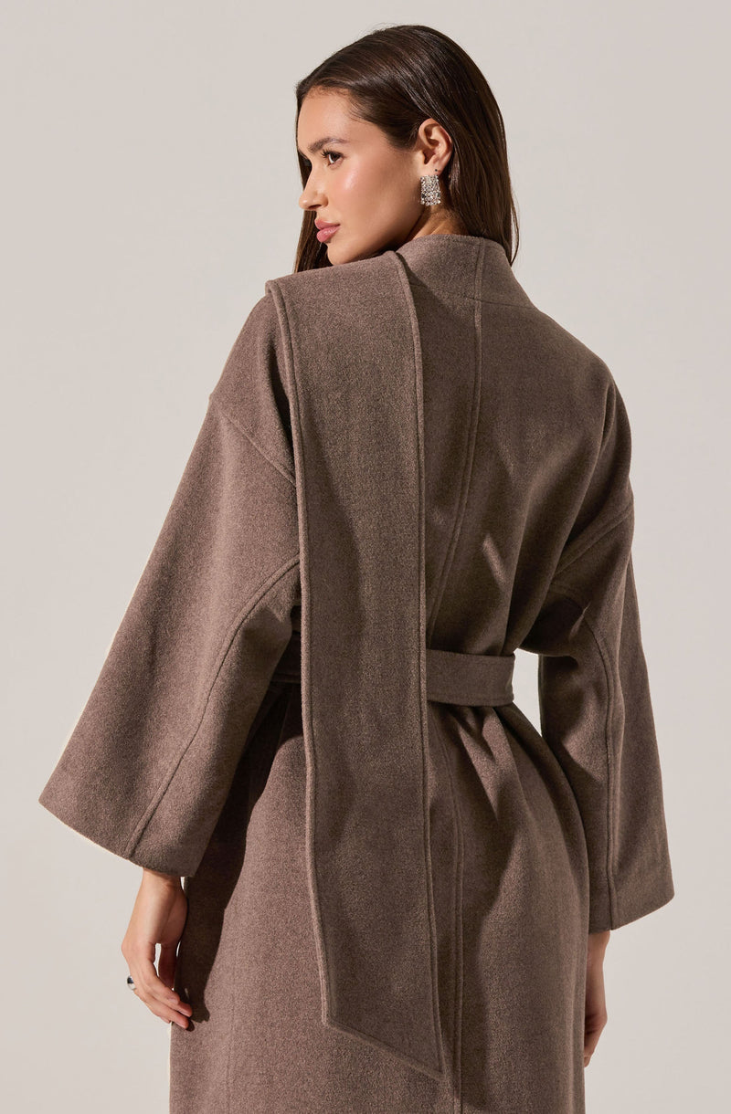 A woman is wearing a long, taupe-colored coat with wide, three-quarter length sleeves and a matching belt tied around the waist. The coat has a high collar and appears to be made of a heavy, warm fabric. She is accessorized with large, sparkly earrings.