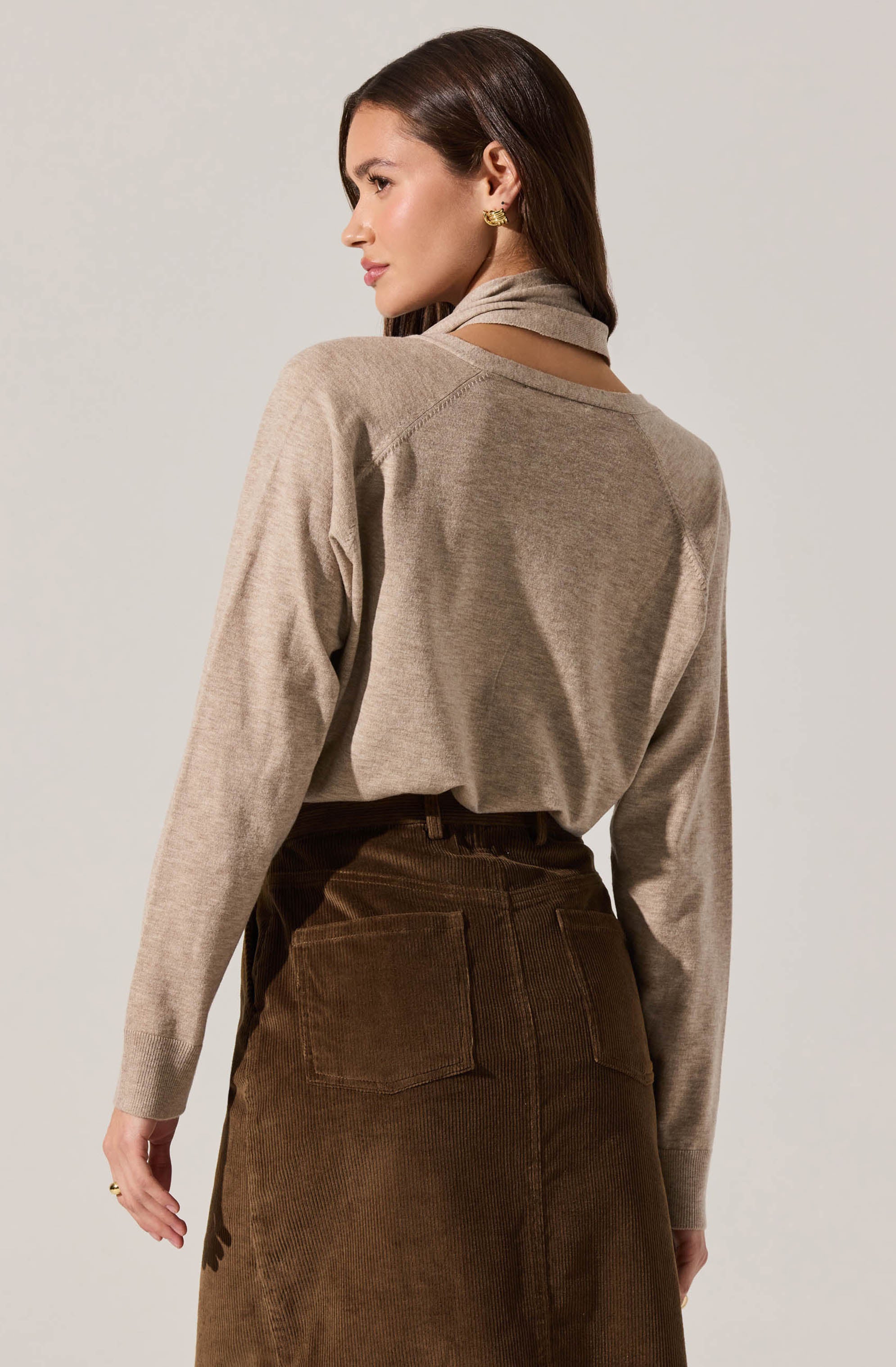 A woman is wearing a beige long-sleeve sweater with a high collar and a brown corduroy skirt with large back pockets. The sweater appears soft and slightly loose-fitting, while the skirt has a high waistband. The woman has straight, dark brown hair and is wearing gold hoop earrings.
