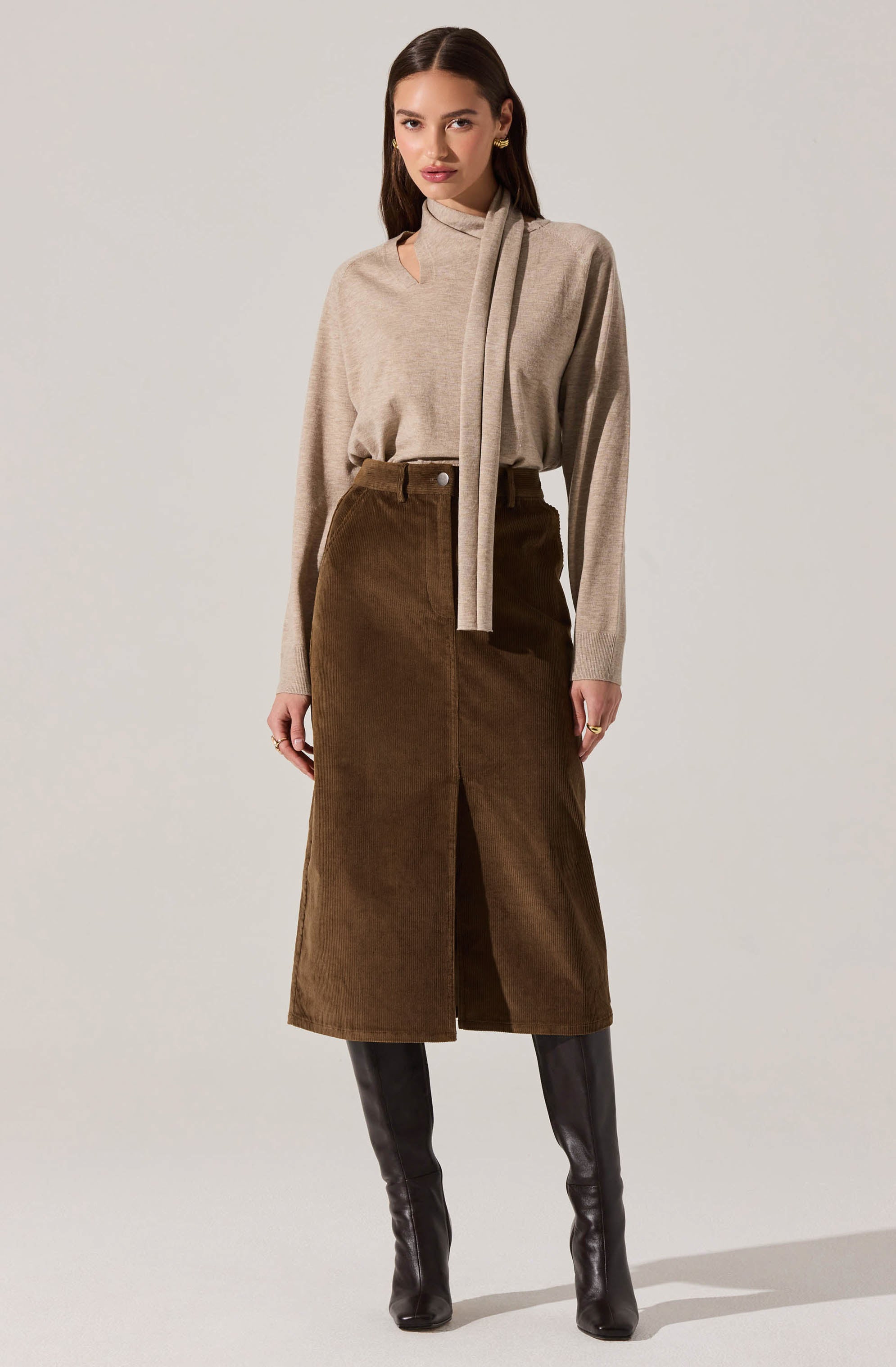 A woman is wearing a beige long-sleeve sweater with a high neckline and a matching scarf draped around her neck. She pairs it with a high-waisted, brown, corduroy maxi skirt that has a front slit and button closure at the waist. She completes the outfit with tall black leather boots.